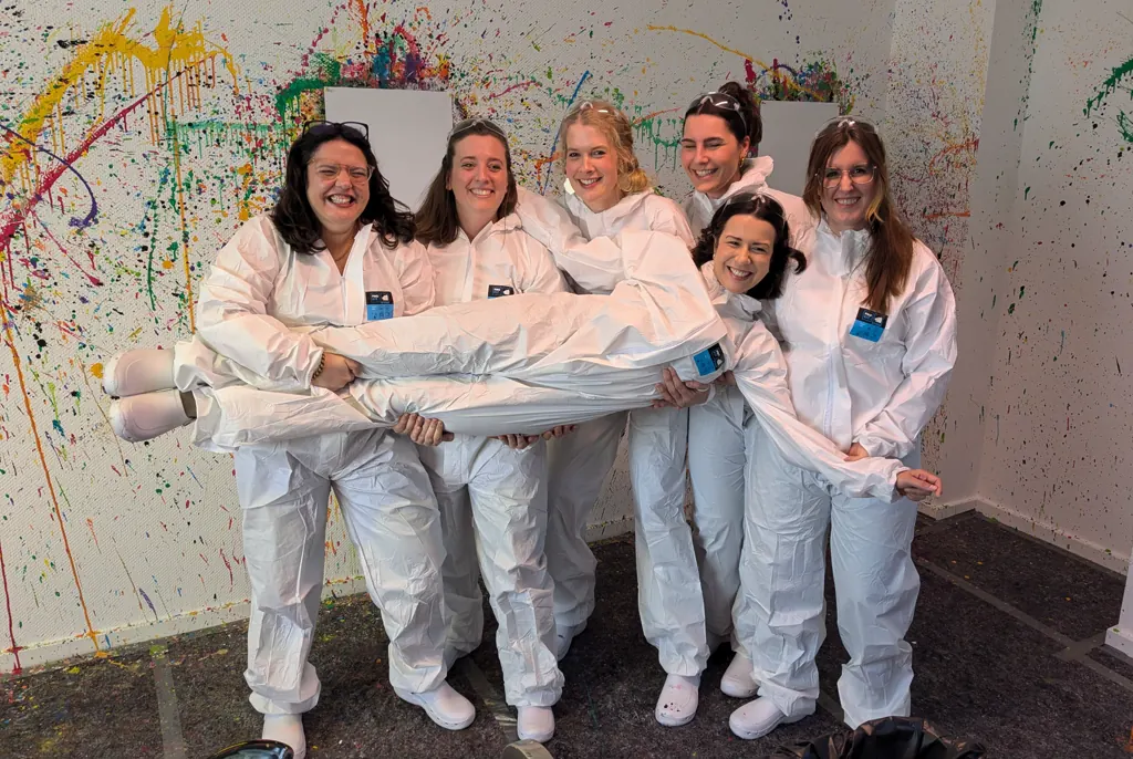 Sechs Frauen in weißen Overalls lachen in die Kamera, eine von ihnen wird quer auf den Armen der anderen getragen, dahinter eine farbbespritzte Wand