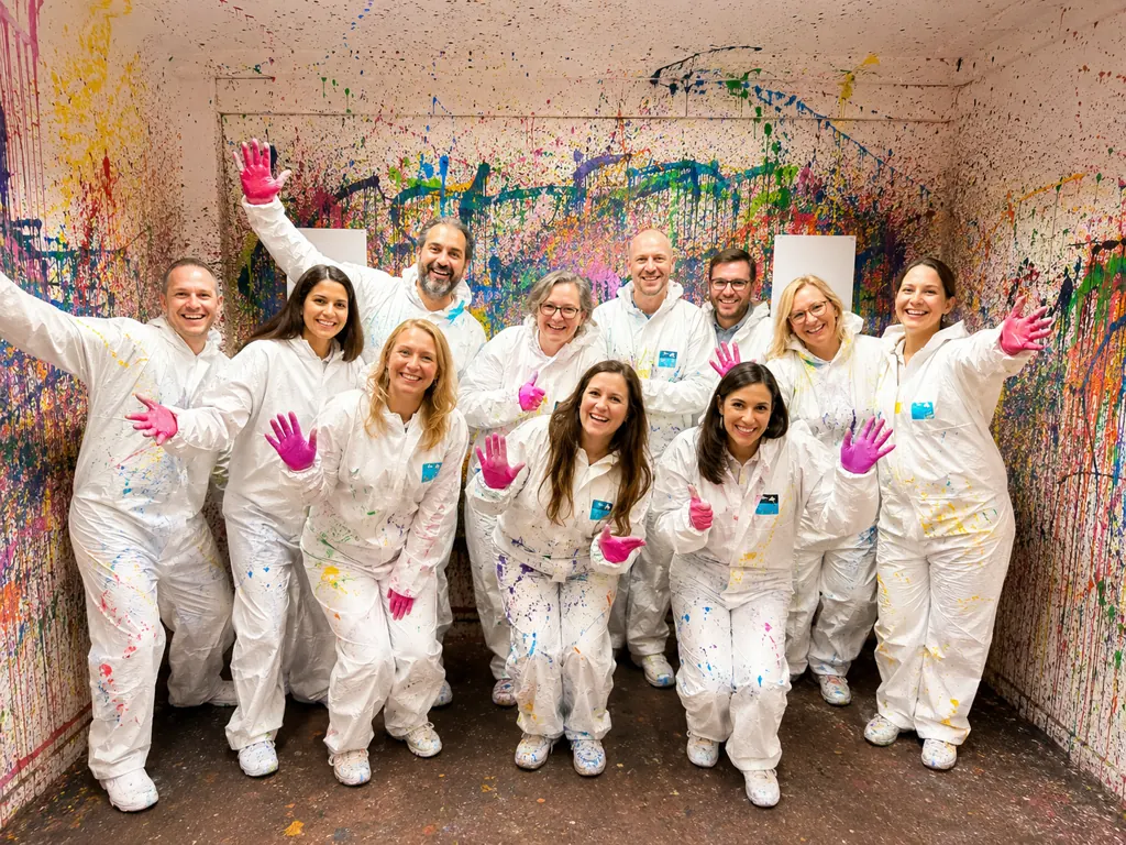 Eine Gruppe in weißen Overalls und pinken Handschuhen posiert mit erhobenen Händen in einem Raum, dessen Wände mit bunter Farbe bespritzt sind