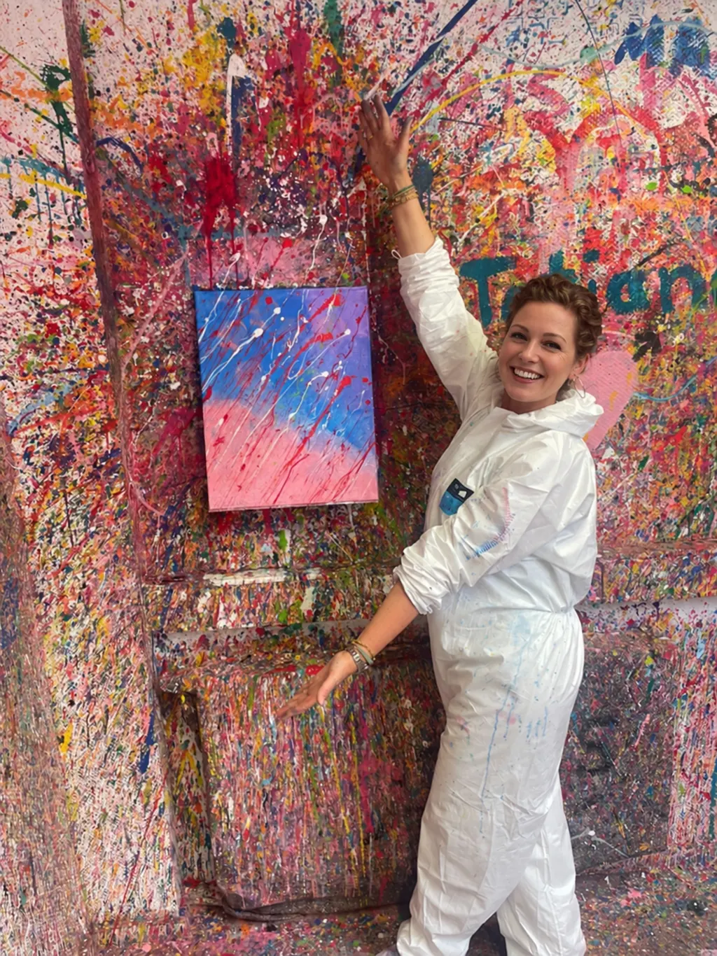 Eine Frau im Overall zeigt stolz ihr fertiges Bild in Rosa und Blau an der bespritzten Wand
