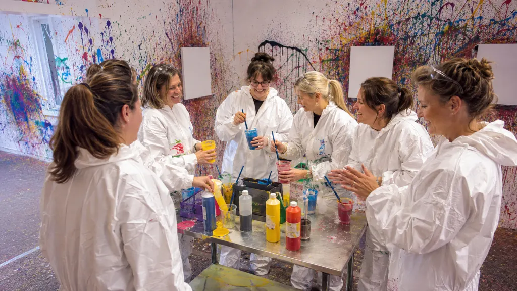 Sechs Frauen in weißen Overalls rühren am Farbtisch bunte Farbe an, dahinter leere Leinwände an der bespritzten Wand
