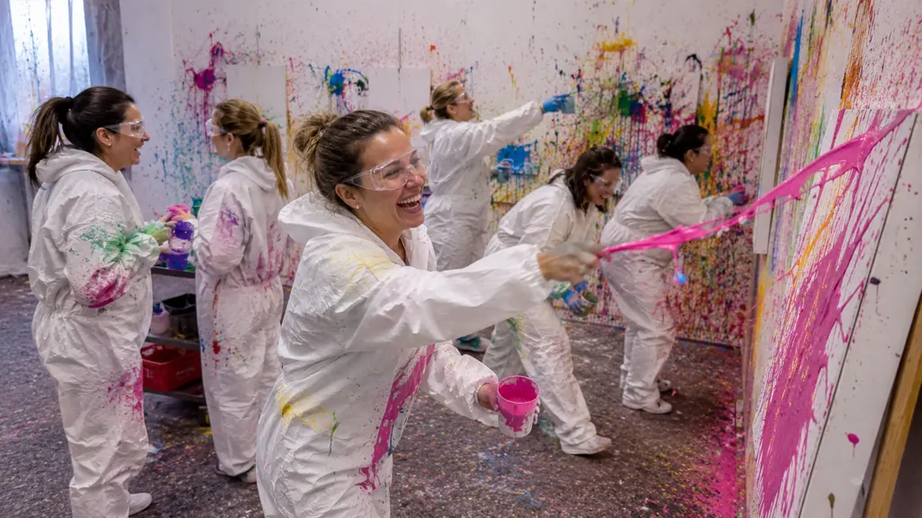 Eine Frau im weißen Overall schleudert pinke Farbe auf die Leinwand, hinter ihr lachen Freundinnen vor der bunt bespritzten Wand