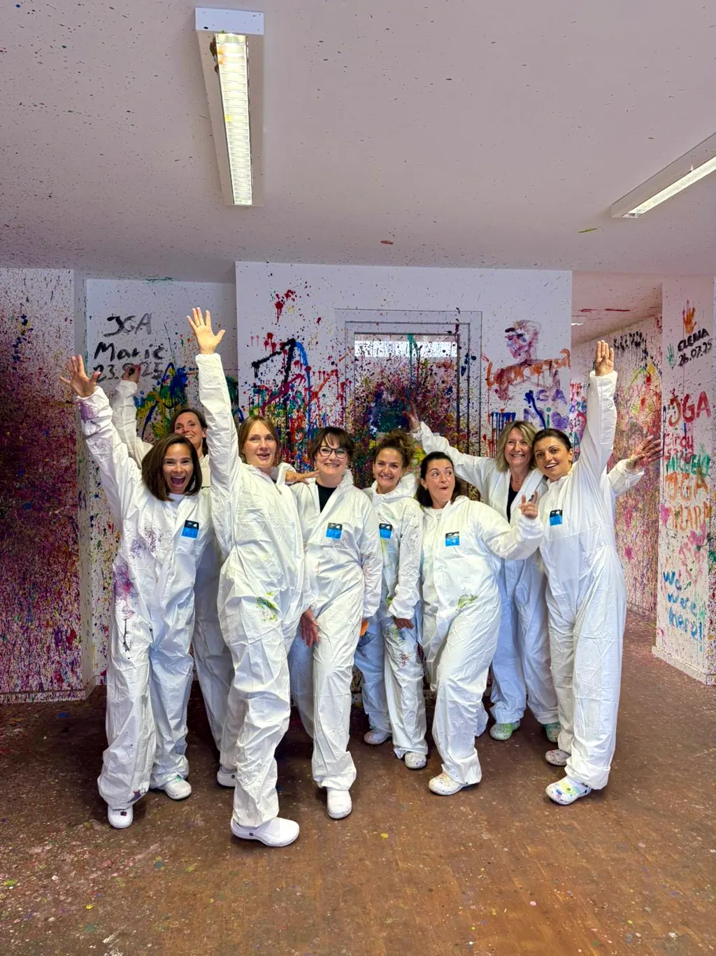 JGA-Gruppe in weißen Overalls jubelt mit erhobenen Armen vor einer Wand voller Namen und Farbe