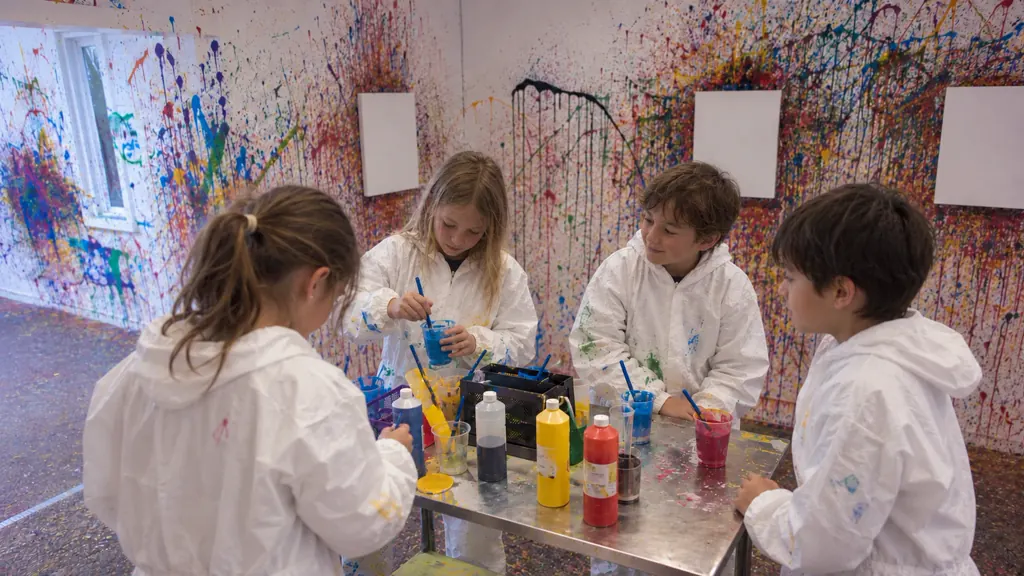 Vier Kinder in weißen Overalls stehen am Farbtisch und rühren blaue Farbe an, dahinter leere Leinwände an der bunt bespritzten Wand
