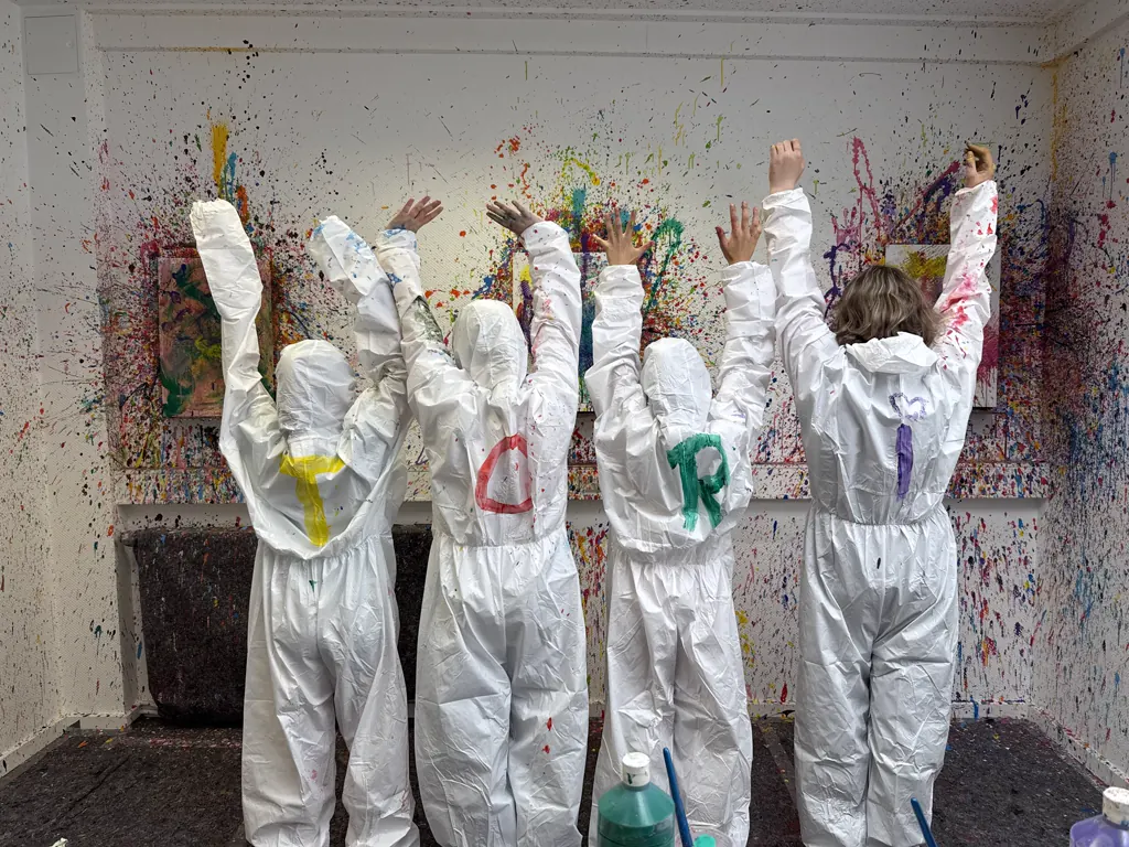 Vier Kinder in weißen Overalls von hinten, die Arme hochgestreckt, auf den Rücken gemalte Buchstaben, vor ihnen die bespritzte Wand mit Leinwänden